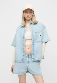 Νεαρή γυναίκα που φοράει ανοιχτόχρωμο κοντό τζιν σορτς, φαρδιά κοντομάνικη τζιν πουκάμισο πάνω από μπεζ crop top με μακριά μανίκια, λευκό καπέλο LA.