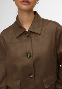 Veste en daim marron avec col, deux boutons et poches. Texture lisse, coupe ajustée et couleur neutre. Présente un design minimaliste.