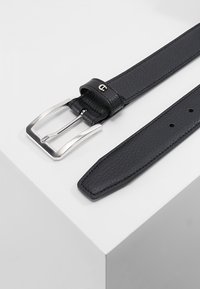 Cinturón de cuero negro con acabado texturizado, hebilla plateada y un lazo, diseñado para un ajuste seguro. Cuenta con varios agujeros ajustables.