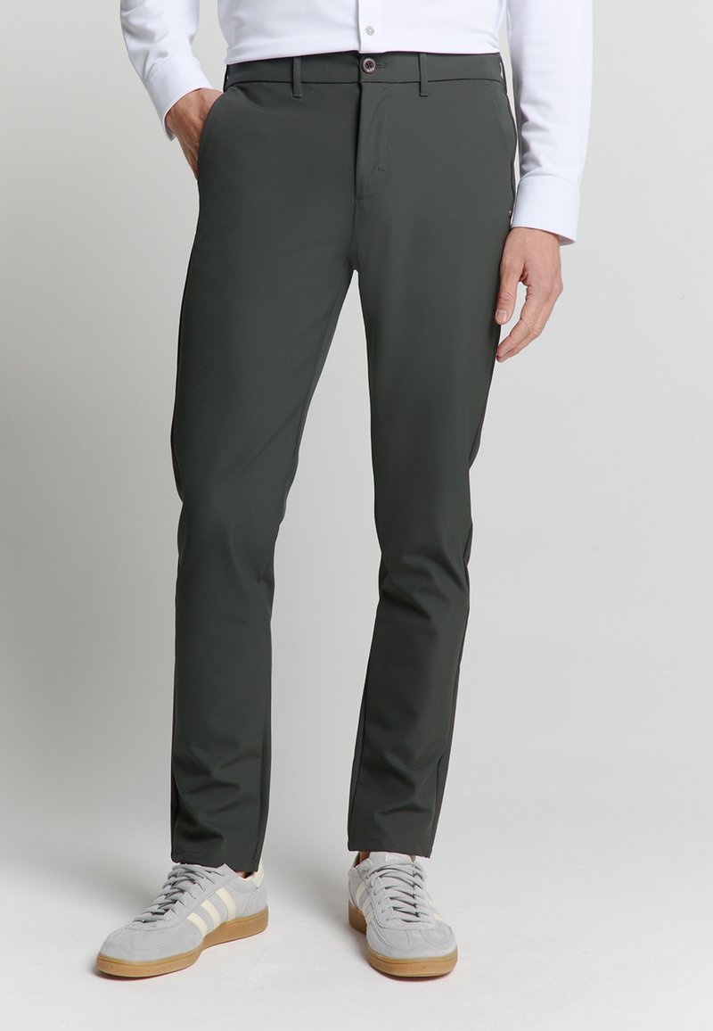 Donkergroene broek van gladde stof, met een slanke pasvorm, steekzakken en een knoopsluiting, gecombineerd met grijze sneakers.