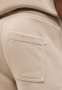 Κοντινή λήψη στην τσέπη μπεζ φόρμας με μια μικρή λευκή ετικέτα με την επιγραφή "LINDSEY LOH".