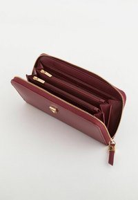 Portefeuille en cuir bordeaux avec fermeture éclair dorée, comprenant plusieurs compartiments et un extérieur texturé. Détail de fermeture à l'avant.