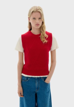 Rode gebreide mouwloze trui met een textured ruitpatroon, gecombineerd met een witte T-shirt met korte mouwen en blauwe wijde jeans.