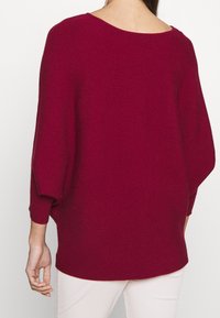 Femme portant un pull ample à manches longues, côtelé et de couleur rouge foncé, avec un large col, accompagné d'un pantalon clair, vue de dos.