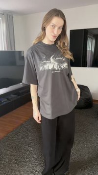 Dunkelgraues, übergroßes T-Shirt mit einem weißen Grafikdesign, das Bäume und einen Nachthimmel darstellt, kombiniert mit schwarzen, weiten Hosen.