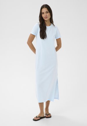 Femeie cu păr lung și închis la culoare, purtând o rochie maxi albastru deschis cu mâneci scurte și șlapi negri, stând cu mâinile la spate.