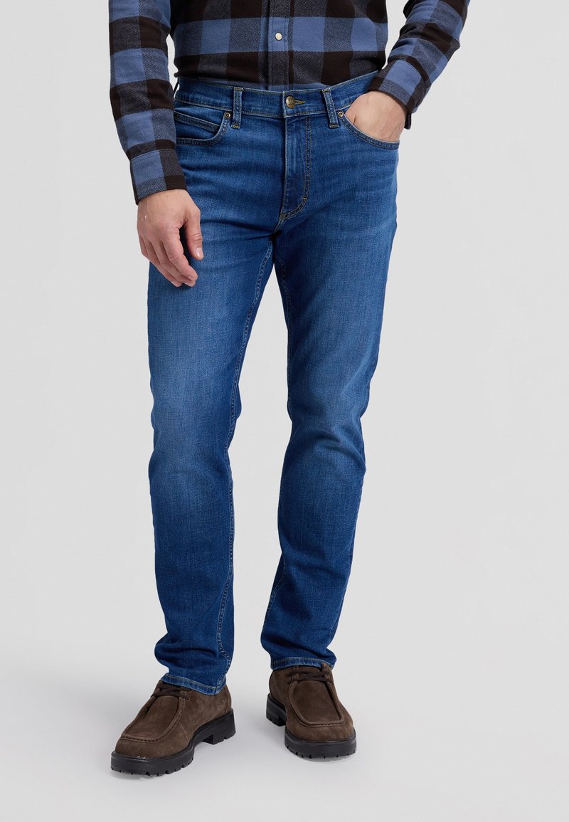 Homme portant un jean bleu, des chaussures en daim marron et une chemise à carreaux noire et bleue, debout avec une main dans la poche.