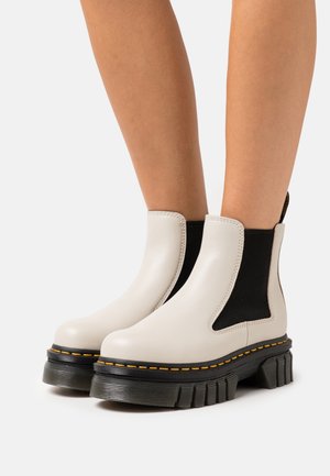 Botas Chelsea de cuero beige con paneles elásticos laterales negros, suela de goma negra gruesa y costuras amarillas a lo largo de los bordes de la suela.