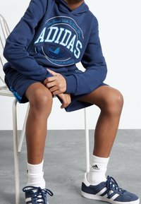 Kind zittend op witte stoel, gekleed in blauwe Adidas hoodie, bijpassende shorts, witte Adidas sokken en blauwe Adidas sneakers op de gray vloer.