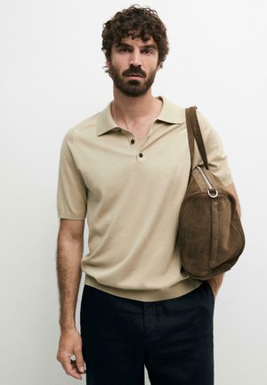 Mann mit lockigem Haar und Bart, trägt beiges kurzärmeliges Poloshirt und schwarze Hose, trägt braune Wildleder-Umhängetasche vor weißem Hintergrund.