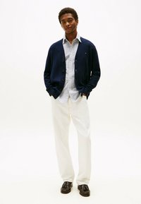 Marineblauer Cardigan, gestreiftes Hemd, cremefarbene Hose und dunkle Schuhe. Der Cardigan hat Knöpfe und lange Ärmel; das Hemd hat einen Kragen.