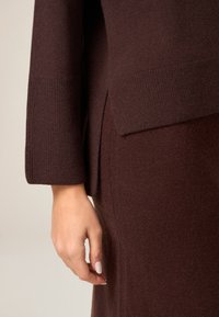 Dunkelbrauner Strickpullover mit gerippten Bündchen und Seitenschlitz. Fokus auf strukturierte Stoff und figurbetonte Silhouette, wobei die auf dem Kleidungsstück ruhende Hand hervorgehoben wird.