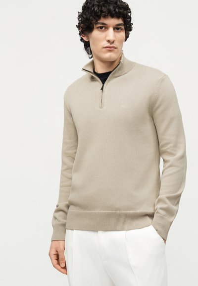 Noor mees lokkis tumedate juustega, kes kannab beeži neljandikul tõmblukuga kampsunit ja valgeid pükse, seisab ühtlase heleda tausta ees.