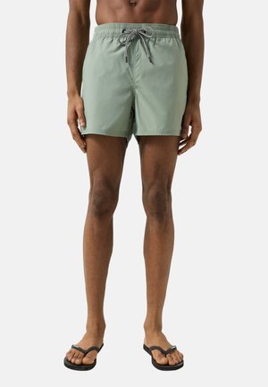 Mand iført lysegrønne badebukser med snor og sorte flipflops, stående foran en ensartet lys baggrund.