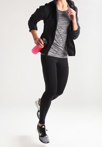 Leggings deportivas negras, camiseta gris a rayas, chaqueta negra con cremallera y zapatillas con estampado. Sosteniendo una botella de agua rosa. Pose activa.