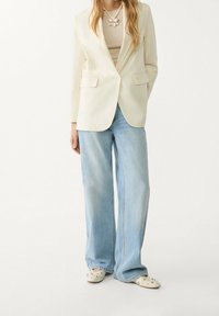 Kremowa marynarka z jednym guzikiem, zestawiona z jasnoniebieskimi spodniami o szerokich nogawkach. Noszona z beżową bluzką i białymi chodakami z ćwiekami.