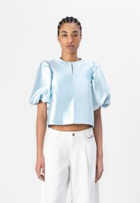 Lyseblå sateng topp med korte ermer og puff-ermer, rund hals og frontslitt. Den har en cropped design og er kombinert med hvite bukser.