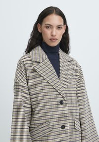 Donna con lunghi capelli scuri indossa un blazer oversize beige e blu a quadri sopra un dolcevita blu, posando contro uno sfondo chiaro e semplice.