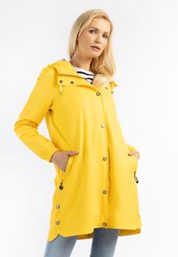 Cappotto impermeabile giallo con cappuccio, accessori metallici argentati, zip frontale, bottoni a pressione e due tasche laterali. Dettaglio delle cuciture all'orlo.
