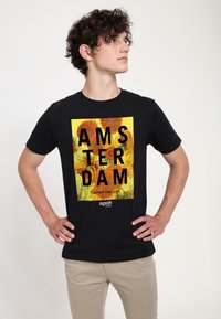 Černé tričko s žlutým obdélníkovým grafickým motivem, na kterém je tučně napsáno "AMSTERDAM" a doplněno květinovým designem inspirovaným Van Goghem.