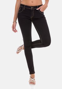 Zwarte denim skinny jeans met oranje stiksels, hoge taille, twee voortassen en een knoopsluiting. Draagt met witte sandaaltjes met bandjes.