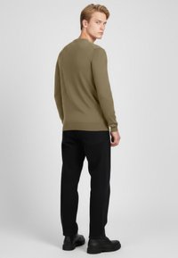 Mladý muž stojící zády, oblečený v olivově zelené košili s dlouhými rukávy, černých kalhotách a černých botách, na jednobarevném šedém pozadí.