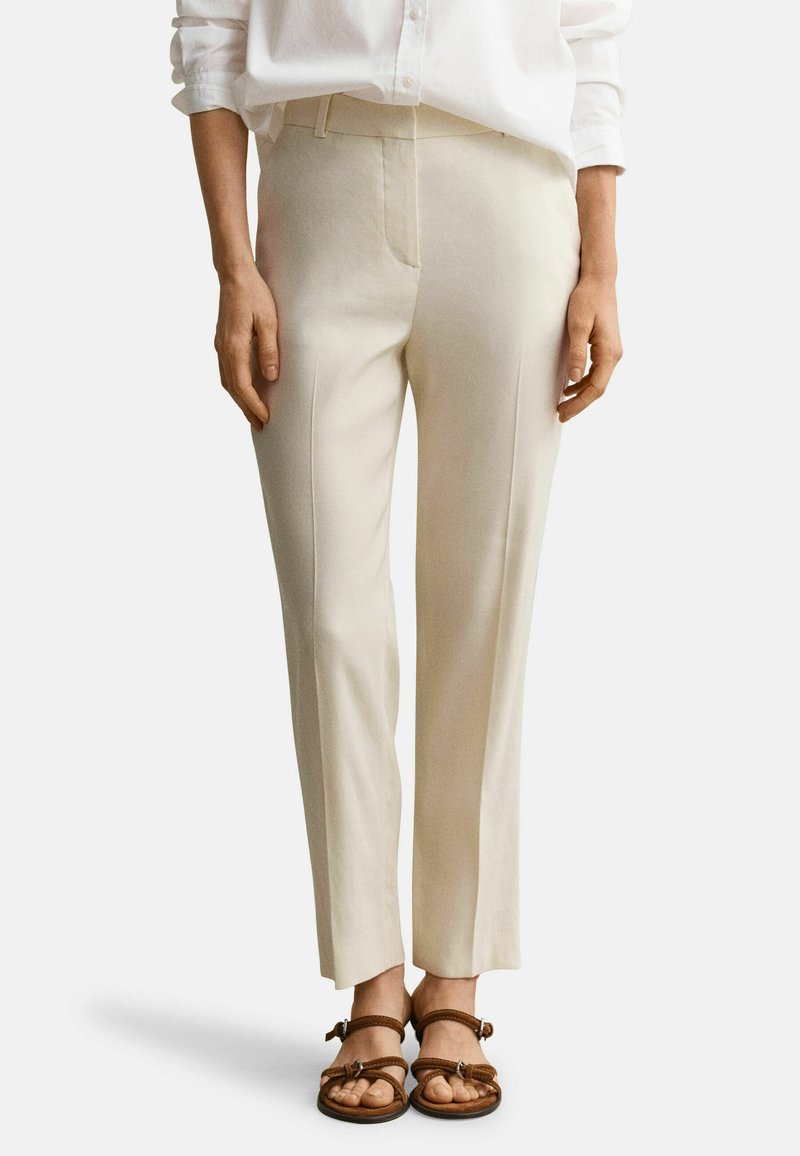 Vrouw draagt beige op maat gemaakte broek en bruine sandalen met bandjes, staat met handen ontspannen langs haar zij tegen een witte achtergrond.