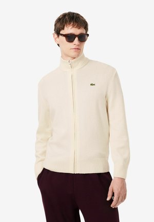 Mann mit dunkler Sonnenbrille, cremefarbenem Reißverschluss-Pullover mit kleinem grünem Logo und dunklen Hosen, steht mit einer Hand in der Tasche.