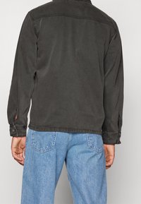 Man som bär en svart denimjacka och ljusblå jeans, visad från ryggen mot en enkel ljus bakgrund.