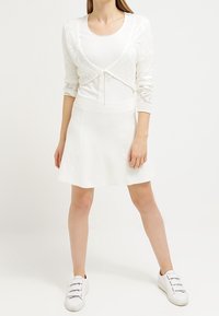 Vit stickad kort cardigan med knytning framtill, kombinerad med en vit figursydd klänning och vita sneakers, med en strukturerad design.