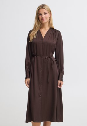 Femme blonde portant une robe longue à manches longues marron foncé boutonnée avec une ceinture à la taille, debout devant un fond clair uni.