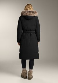 Geacă puffer neagră cu talie marcată, având glugă din blană sintetică și textură matlasată. Poartă cu leggings negri și botine maro.