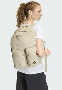 Light beige backpack featuring a textured grid pattern, two front zipper pockets, and adjustable straps. Adidas logo in black on lower right.