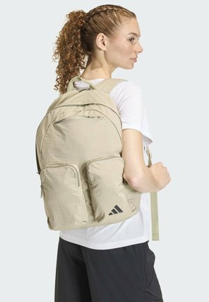 Light beige backpack featuring a textured grid pattern, two front zipper pockets, and adjustable straps. Adidas logo in black on lower right.
