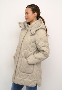 Bežīgs quilted jaka ar kapuci, ar rāvējslēdzēju, sānu kabatām un ģeometrisku rakstu. Audums izskatās gluds un maigs.