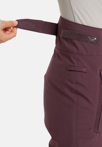 Švestkově červené softshellové kalhoty s nastavitelným suchým zipem v pase, zipovou kapsou a texturovaným akcentem poblíž pásu.