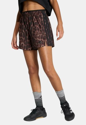 Donna che indossa pantaloncini sportivi marroni con stampa leopardata e strisce laterali nere, scarpe da ginnastica nere e calzini grigi a righe, in piedi su sfondo bianco.