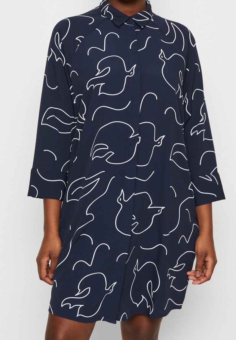 Robe chemise bleu marine avec des contours abstraits d’oiseaux blancs, manches longues et col pointu, portée par une personne à la peau foncée.