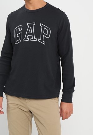 Mand iført en sort langærmet skjorte med "GAP"-logo i hvide bogstaver og beige bukser, stående mod en ensfarvet baggrund.