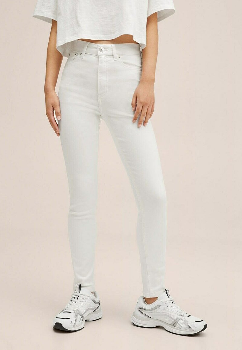 Jean skinny blanc taille haute avec fermeture à bouton, texture lisse et silhouette ajustée, associés à des baskets grises claires.