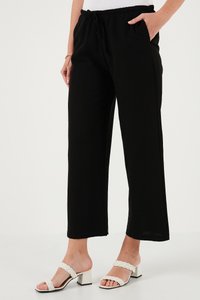 Inimene, kes kannab musta laia säärega pükse elastse vöökohaga, seisab ühe käega taskus ja kannab valgeid punutud rihmadega kontsakingi sandaale.