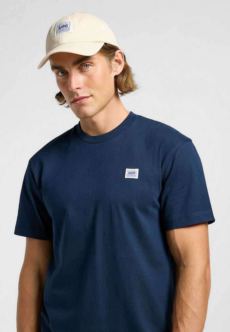 Mörkblå bomullströja med korta ärmar och en liten vit etikett. Beige keps med logotyp, bärs med utsläppt hår. Enkel design, minimala detaljer.