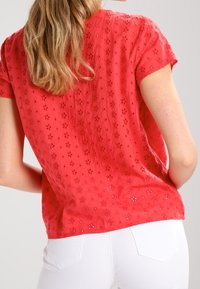 Červená blůza s krátkým rukávem, zdobená květinovou výšivkou a mírně texturovanou tkaninou, kombinovaná s bílými kalhotami.