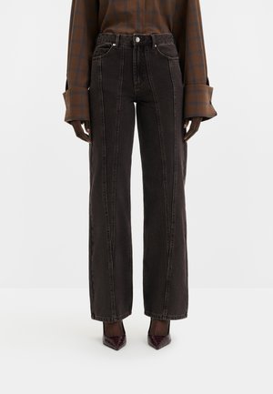 Schwarze High-Waist Jeans mit weitem Bein und vertikalen Nähten, getragen mit einem braunen karierten Hemd und dunklen spitzen Schuhen.