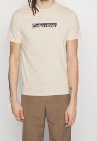 Muž v béžovém tričku Calvin Klein s černým logem a hnědých kalhotách, stojící na jednoduchém bílém pozadí.