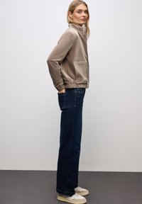 Giacca in corduroy beige con colletto alto, cintura elastica e due tasche frontali, abbinata a jeans dritti blu scuro e sneakers chiare.