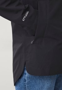 Zwarte, waterdichte jas van gladde stof, voorzien van een zijzak, geborduurde manchetten en een afgeronde zoom. Vergezeld van denim jeans.