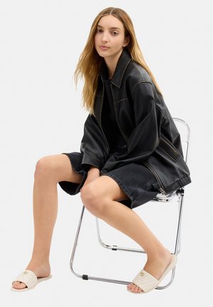 Young woman with long blonde hair wearing a black leather jacket and denim shorts sits on a chrome chair, beige quilted slide sandals on feet.