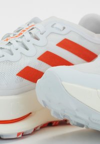 Białe buty sportowe z trzema pomarańczowymi paskami, teksturowana górna część z materiału, wzmocniona pięta oraz biała, wyprofilowana podeszwa z wzorem zwiększającym przyczepność.
