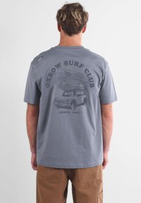 T-shirt gris à manches courtes avec une illustration graphique d'une camionnette portant des planches de surf à l'arrière, et le texte "Oxbow Surf Club" au-dessus.
